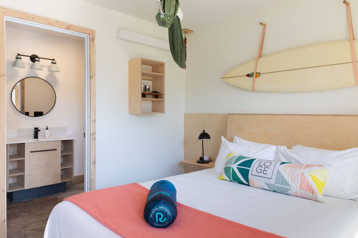 A hotel room with a bed, a bedside table with a lamp, a shelf with toiletries, a hanging surfboard, and a pillow with a geometric pattern.
