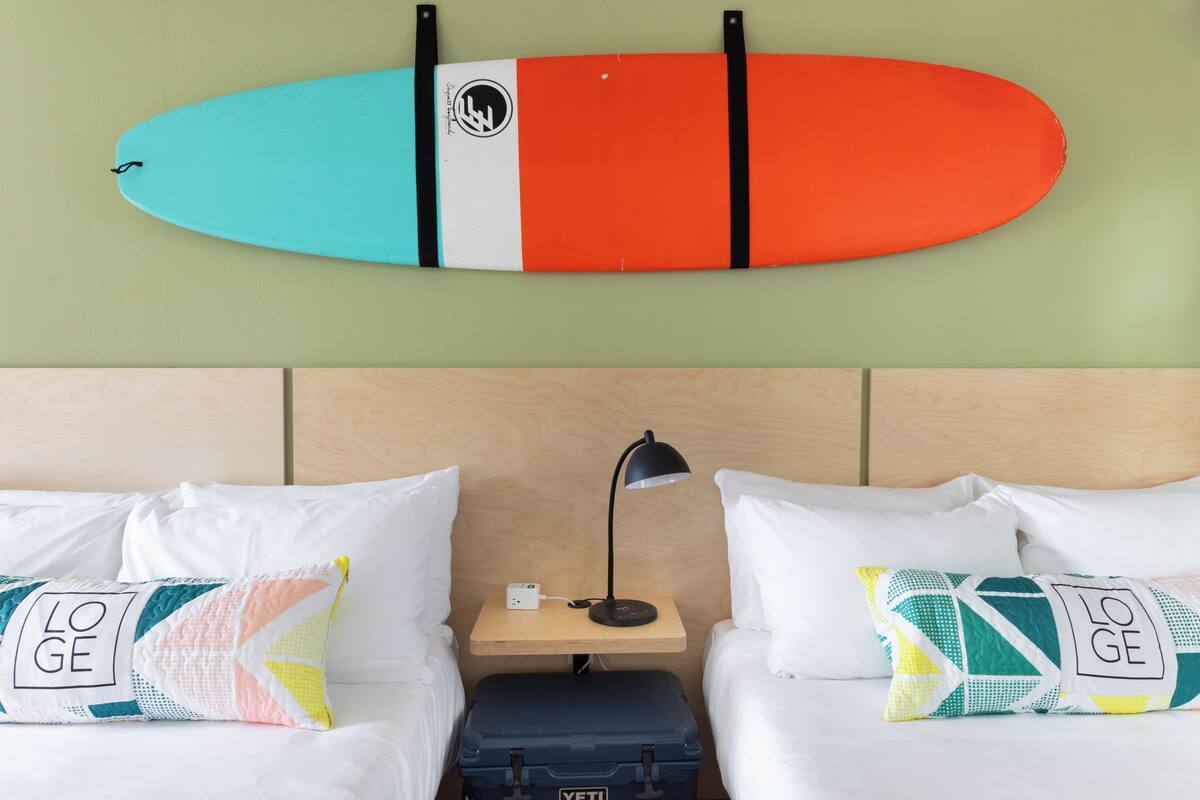 A hotel room with two beds, a nightstand with a lamp, and a surfboard on the wall.