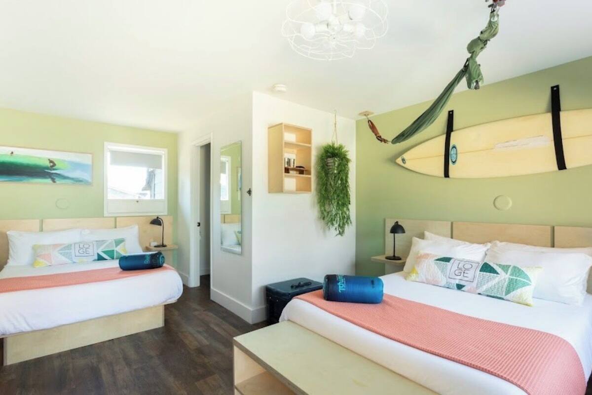A hotel room with two beds, a hammock, a surfboard, and a plant.