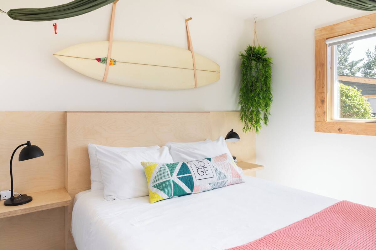 A bedroom with a bed, a hanging surfboard, a potted plant, and a window.