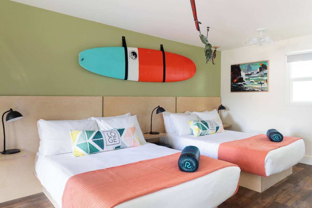 A hotel room with two beds, a surfboard on the wall, a painting, and a ceiling fan.