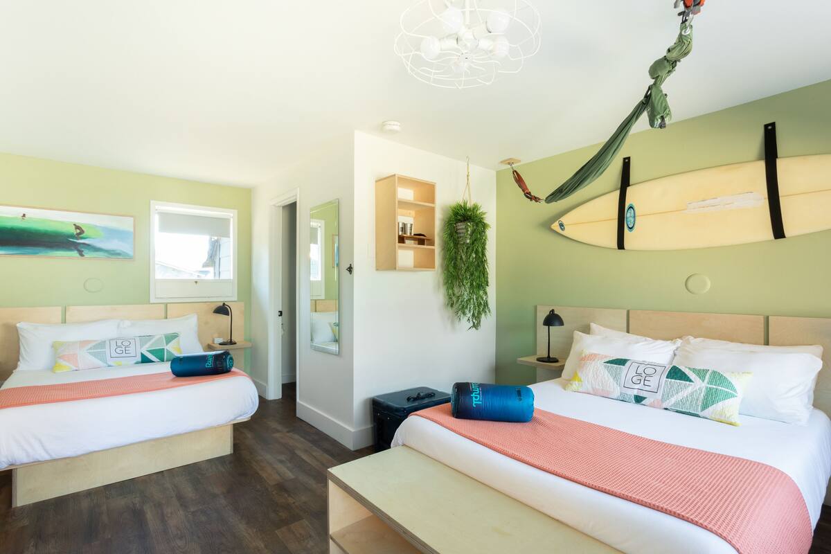 A hotel room with two beds, a hammock, a surfboard, and a small plant.