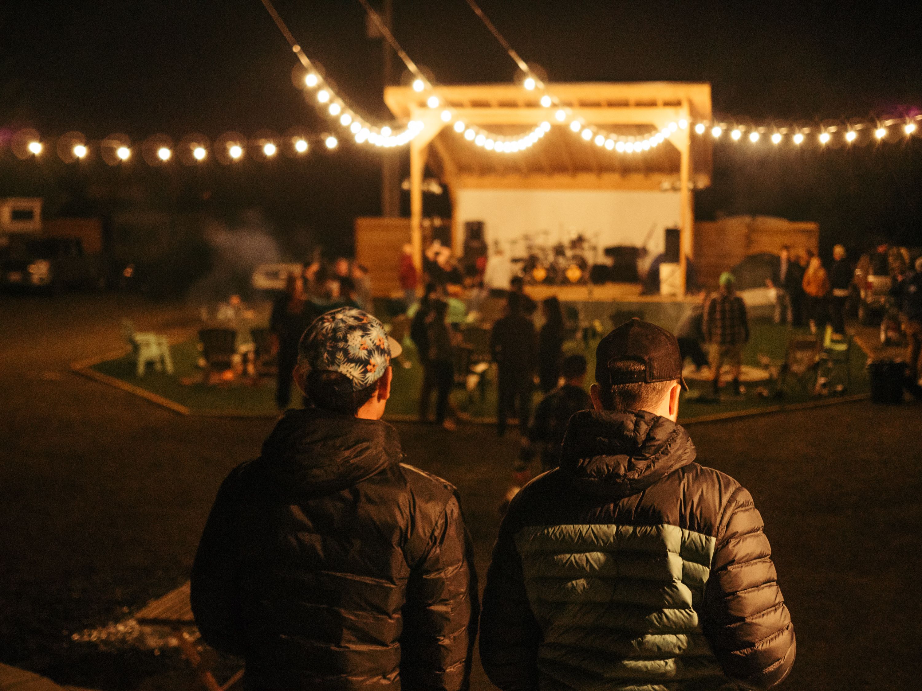 Two people standing in front of a stage with string lights, surrounded by tables and chairs.