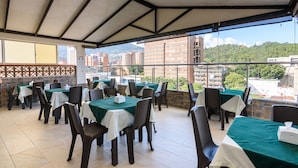 An outdoor dining area with tables and chairs, a tiled floor, and a view of buildings and greenery in the background. - Hotel Med Estadio (Medellín)