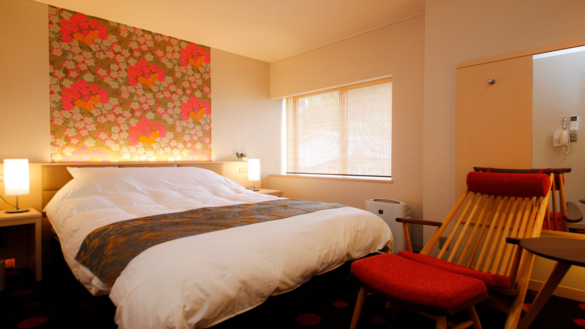 Et hotelværelse med en seng, sengeborde med lamper, et vindue med rullegardiner, en stol og en væg med blomstermønster.