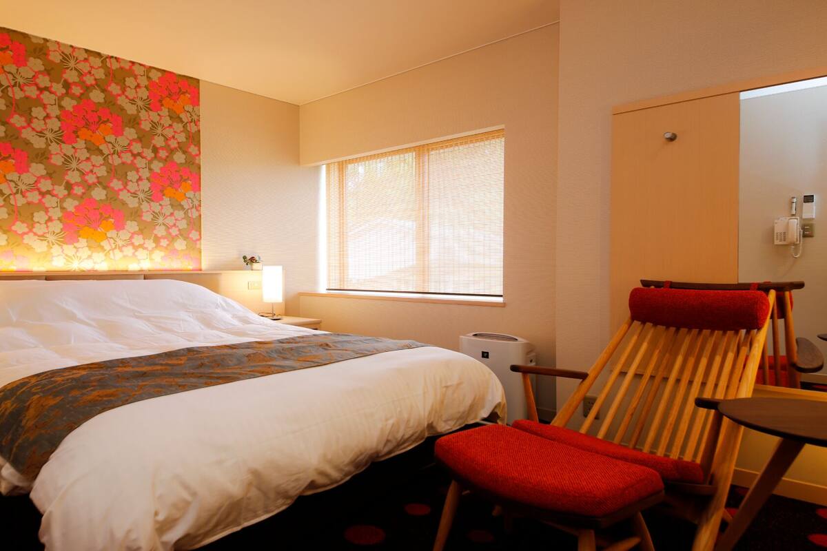 A hotel room with a bed, bedside lamps, a window with blinds, a chair, and a wall with a floral pattern.