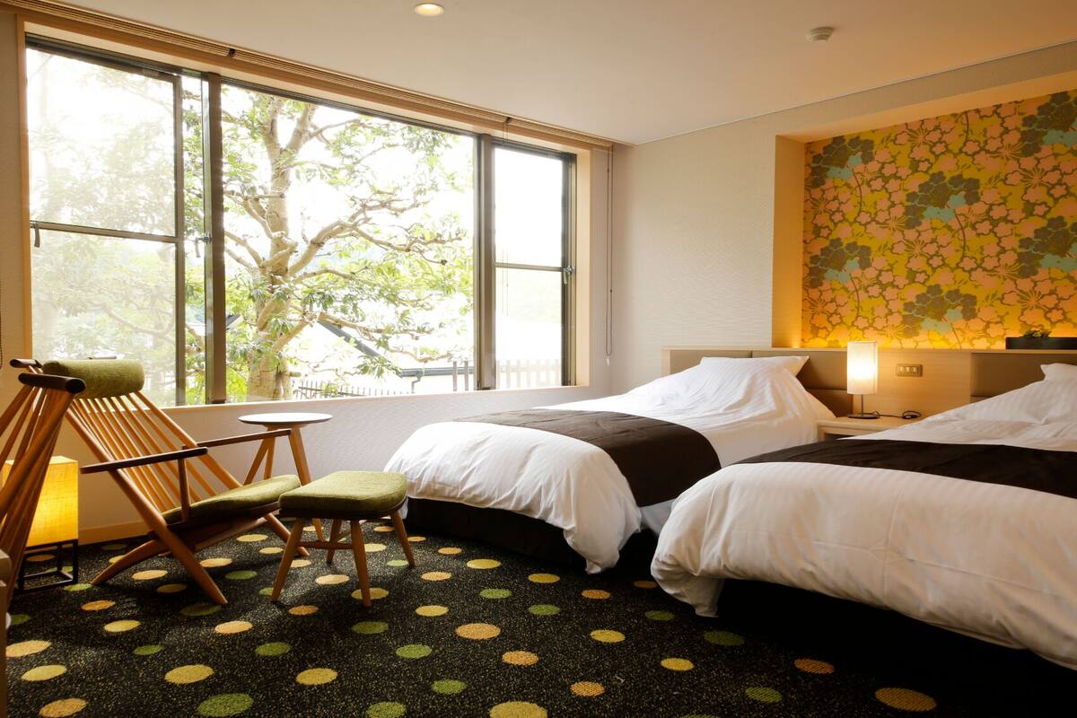 A hotel room with two beds, a chair, a small table, and large windows with a view of trees.