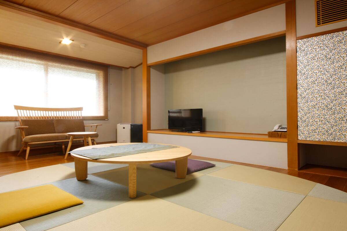 A traditional Japanese room with tatami flooring, a low table, a sofa, a television, and a patterned wall.