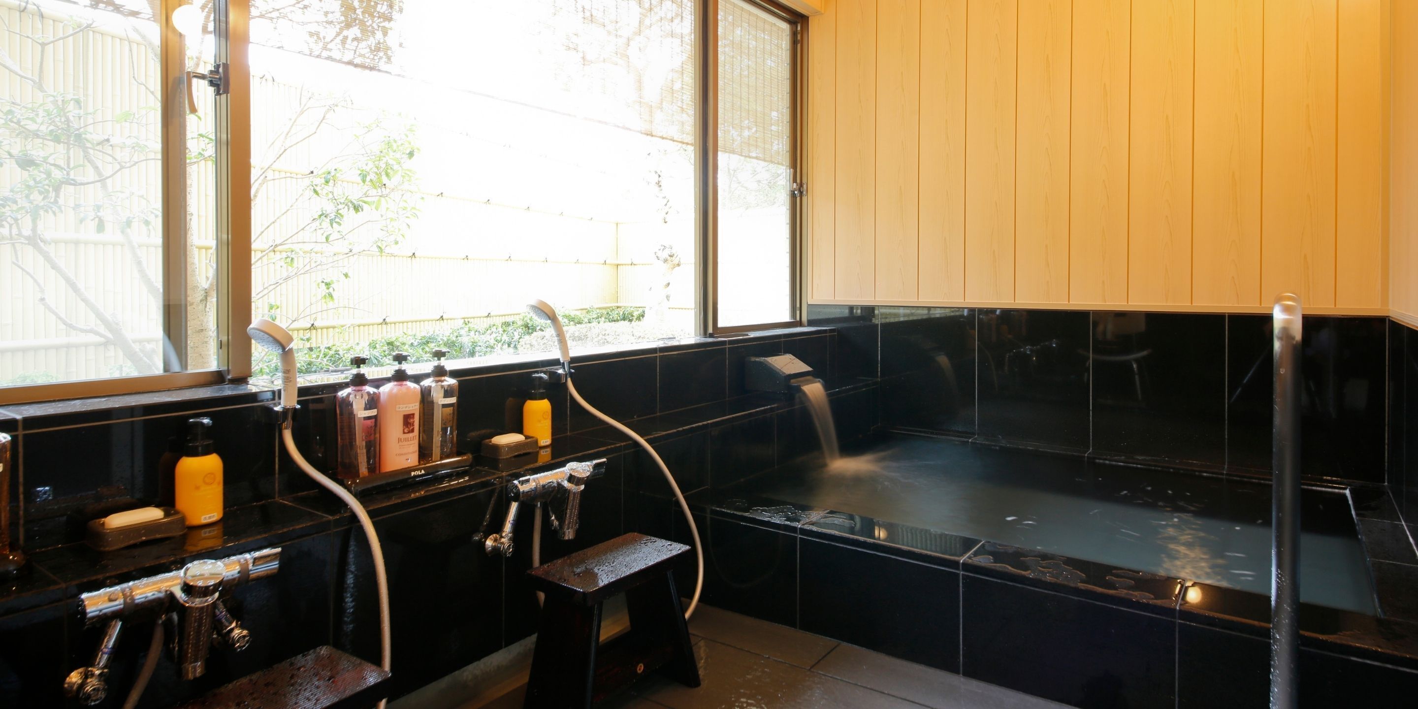 Un baño con una bañera negra, una ventana que da a una valla y árboles, y un lavamanos con distintos artículos de aseo.