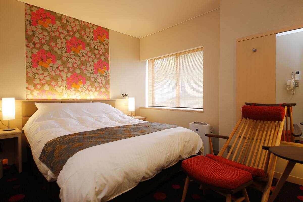 A hotel room with a bed, a chair, a window, and a wall with a floral pattern.