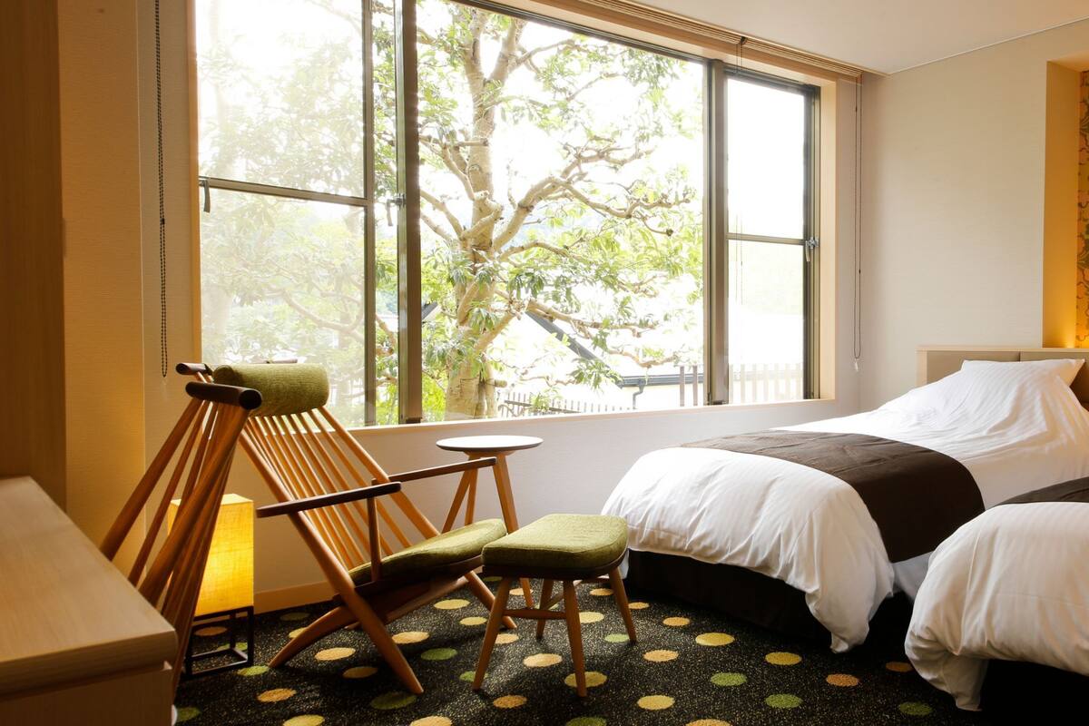 A hotel room with two beds, a small table, a chair, and a large window with a view of trees.