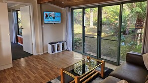 A modern living room with a wooden floor, a glass coffee table, a flat-screen TV mounted on the wall, and a sliding glass door leading to a garden.