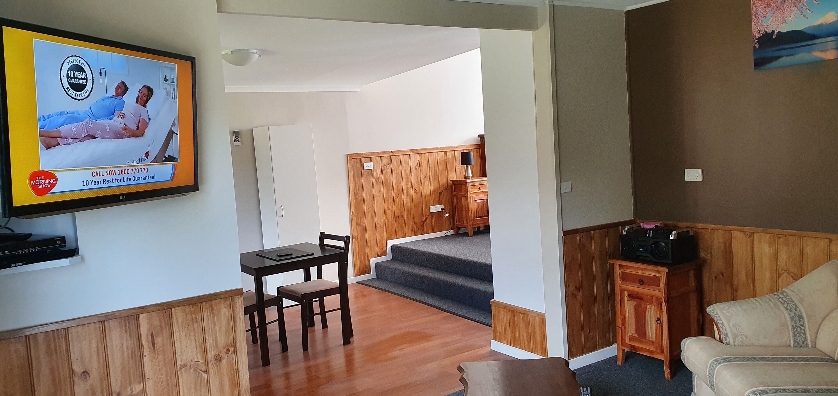 A living room with a TV displaying an advertisement, a dining area with a table and chairs, and a staircase leading to another room.