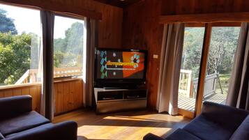 A wooden interior with a television displaying 'MOTORSPORTSNT' and a website URL, wooden flooring, and large windows offering a view of greenery.