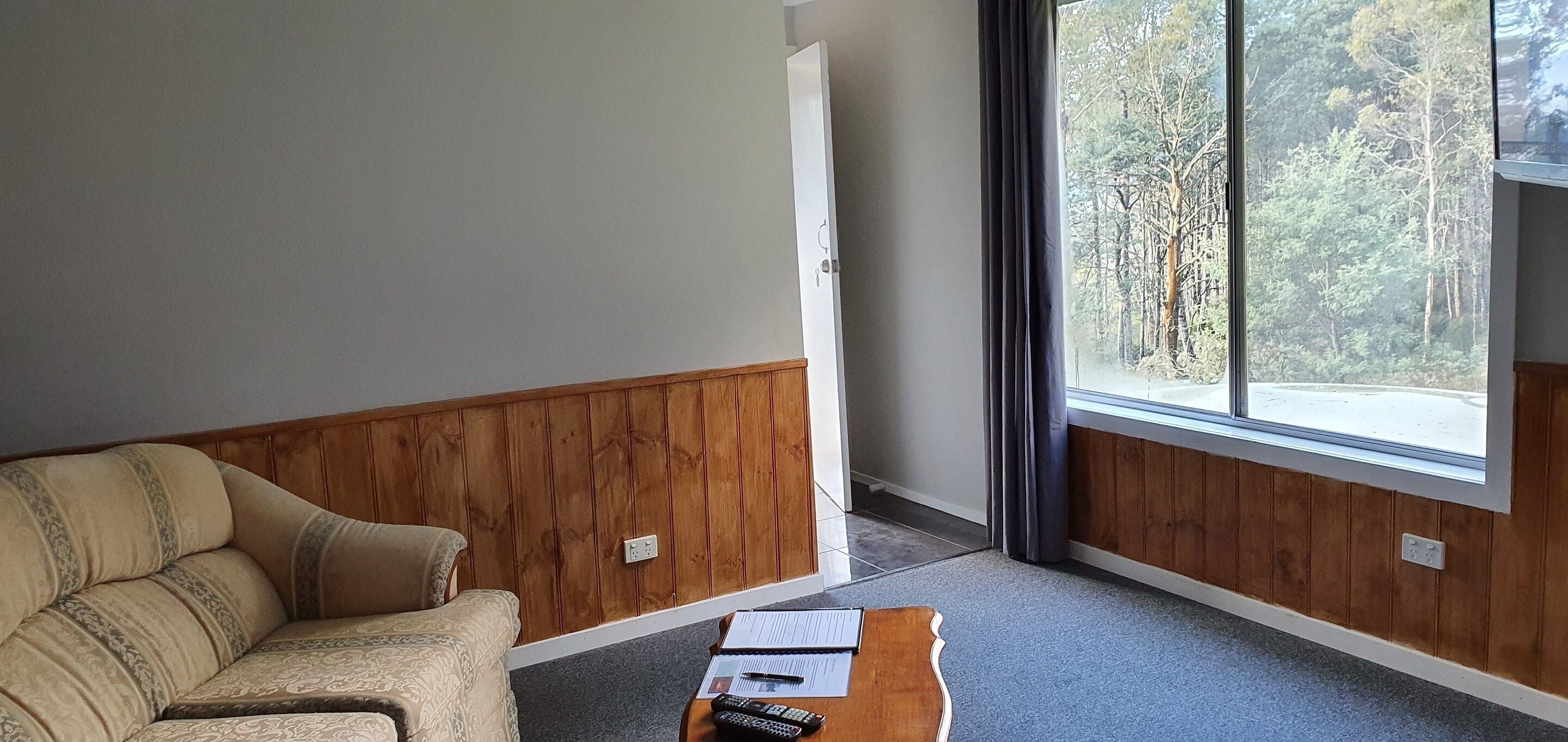 Una habitación con pared de paneles de madera, un sofá beige, una pequeña mesa de madera con papeles y un control remoto, y una ventana con vistas a un bosque.