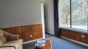 Una habitación con pared de paneles de madera, un sofá beige, una pequeña mesa de madera con papeles y un control remoto, y una ventana con vistas a un bosque.