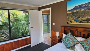 A bedroom with a large window, a bed with white linens, a wooden headboard, and a painting of a mountainous landscape.