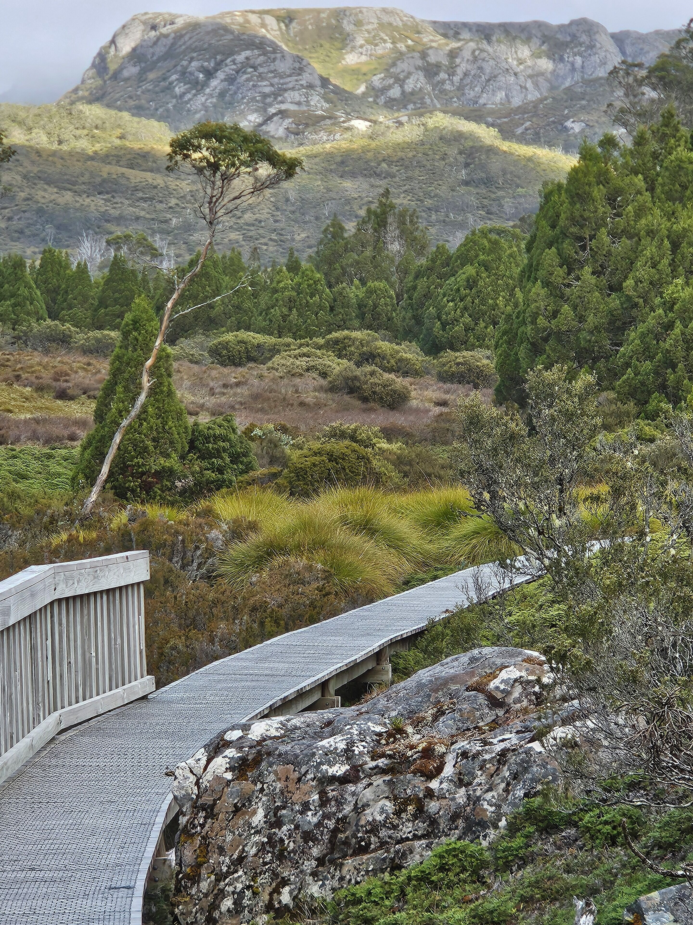 Uma ponte de madeira que leva a uma área rochosa com vegetação escassa e montanhas ao fundo.