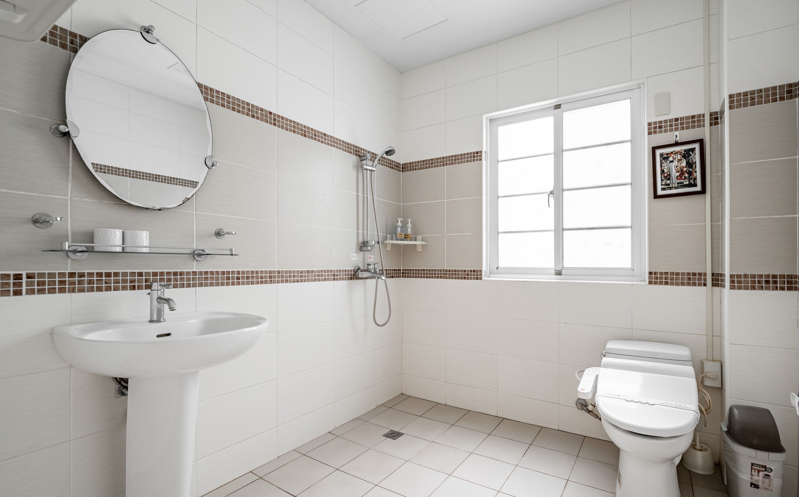 A modern bathroom with a white sink, toilet, and shower. There is a window providing natural light and a mirror above the sink.