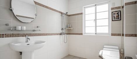 A modern bathroom with a white sink, toilet, and shower. There is a window providing natural light and a mirror above the sink.