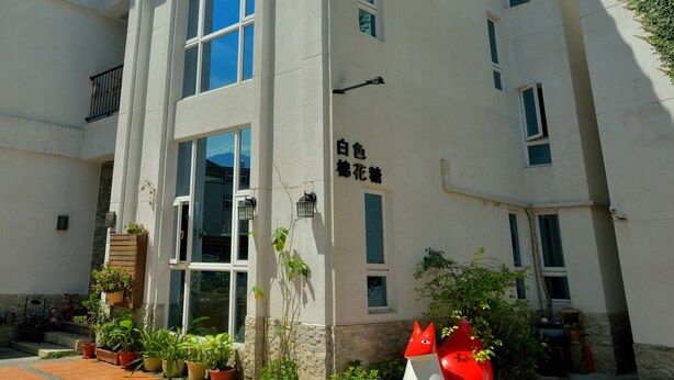 Un edificio de varios pisos con fachada blanca, amplios ventanales y un patio empedrado con plantas en macetas y una llamativa escultura roja.