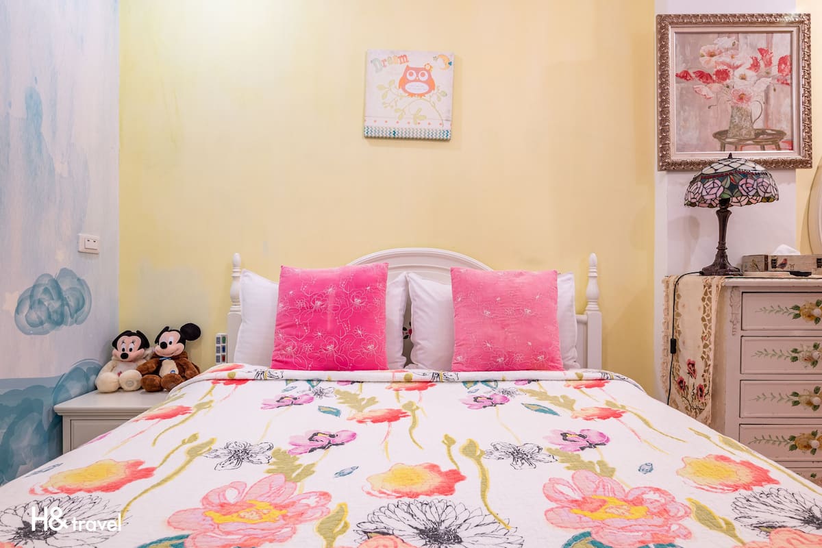 A bedroom with a floral bedspread, two Mickey Mouse plush toys on the bed, and a wall with a framed picture of flowers.