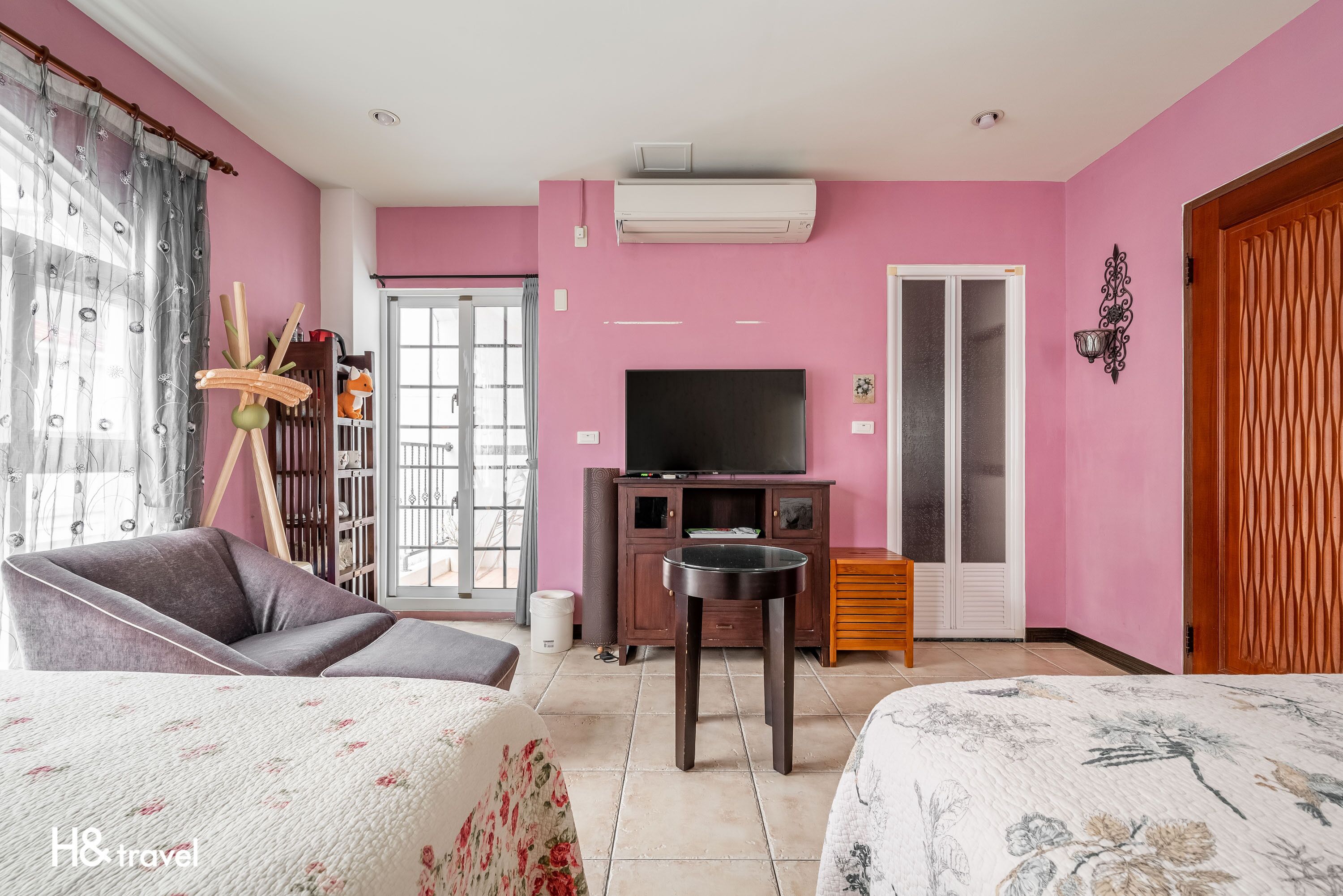 Une pièce aux murs roses, avec un téléviseur, une armoire en bois et un lit recouvert d’une literie à motifs floraux.