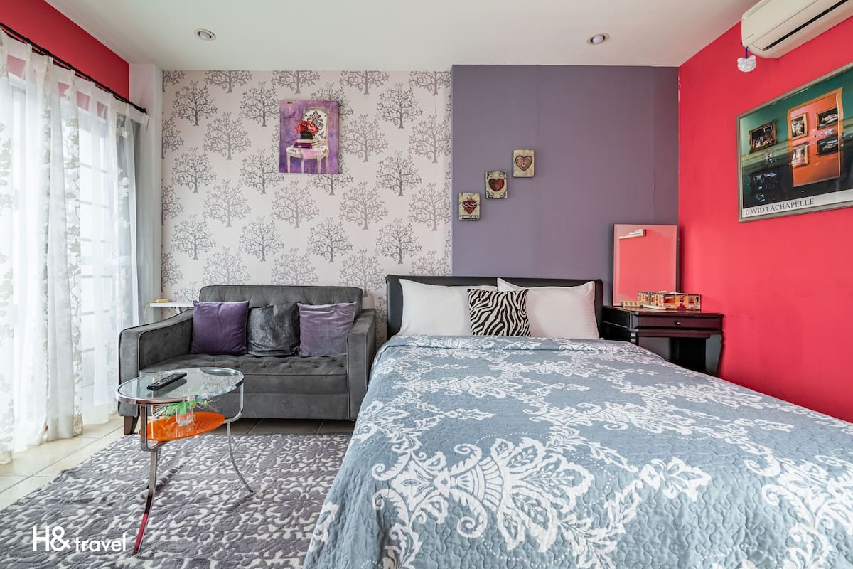 A bedroom with a grey sofa, a glass coffee table, a patterned bedspread, and a red wall with a framed picture.