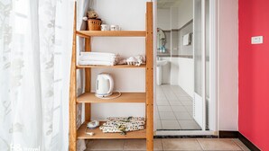Habitación de hotel con repisas de madera que sostienen toallas, una tetera y una cesta. Se ve un baño con lavamanos y espejo a través de una puerta abierta.