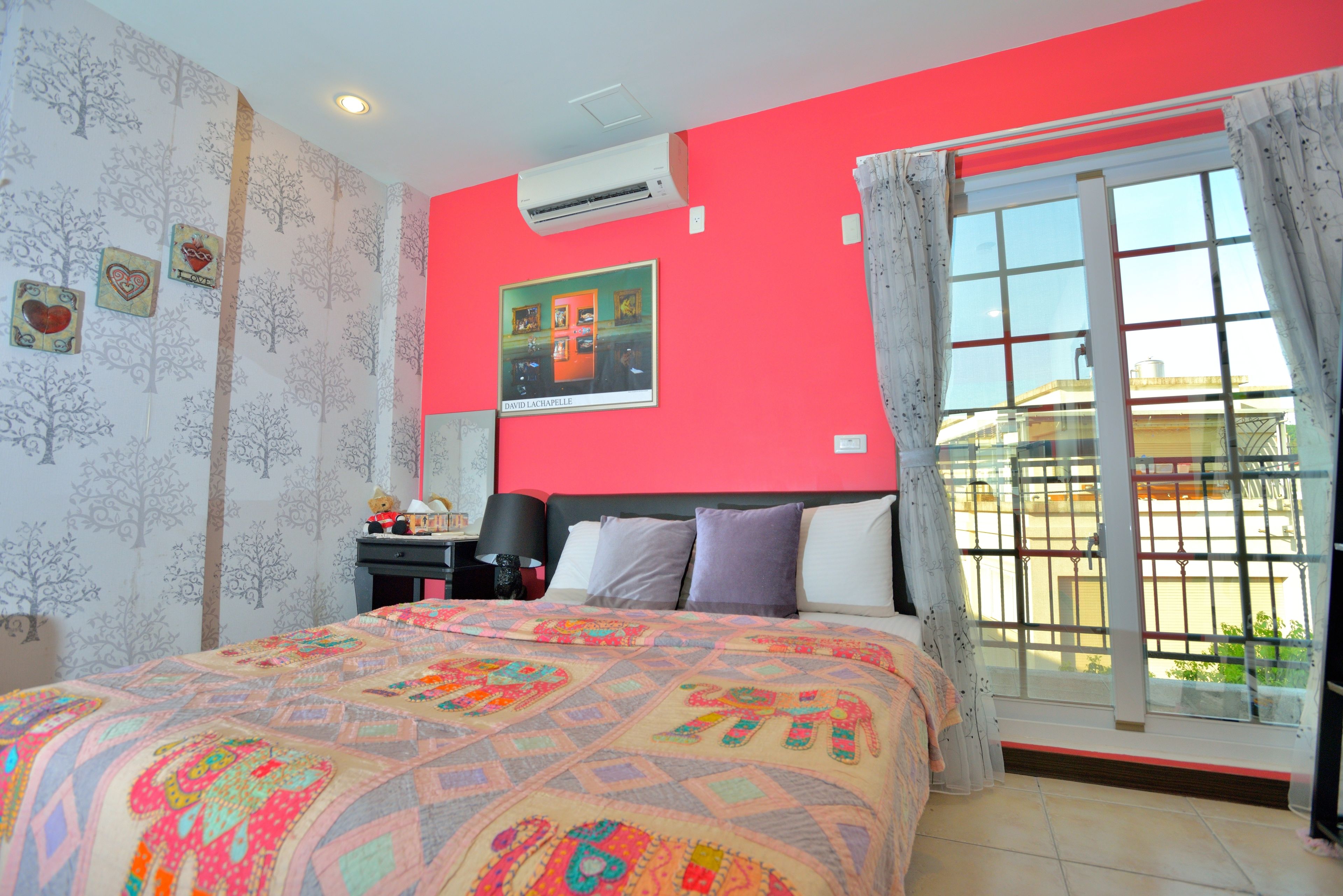 A bedroom with a large bed, a nightstand, a lamp, a window with curtains, and a wall-mounted air conditioner.