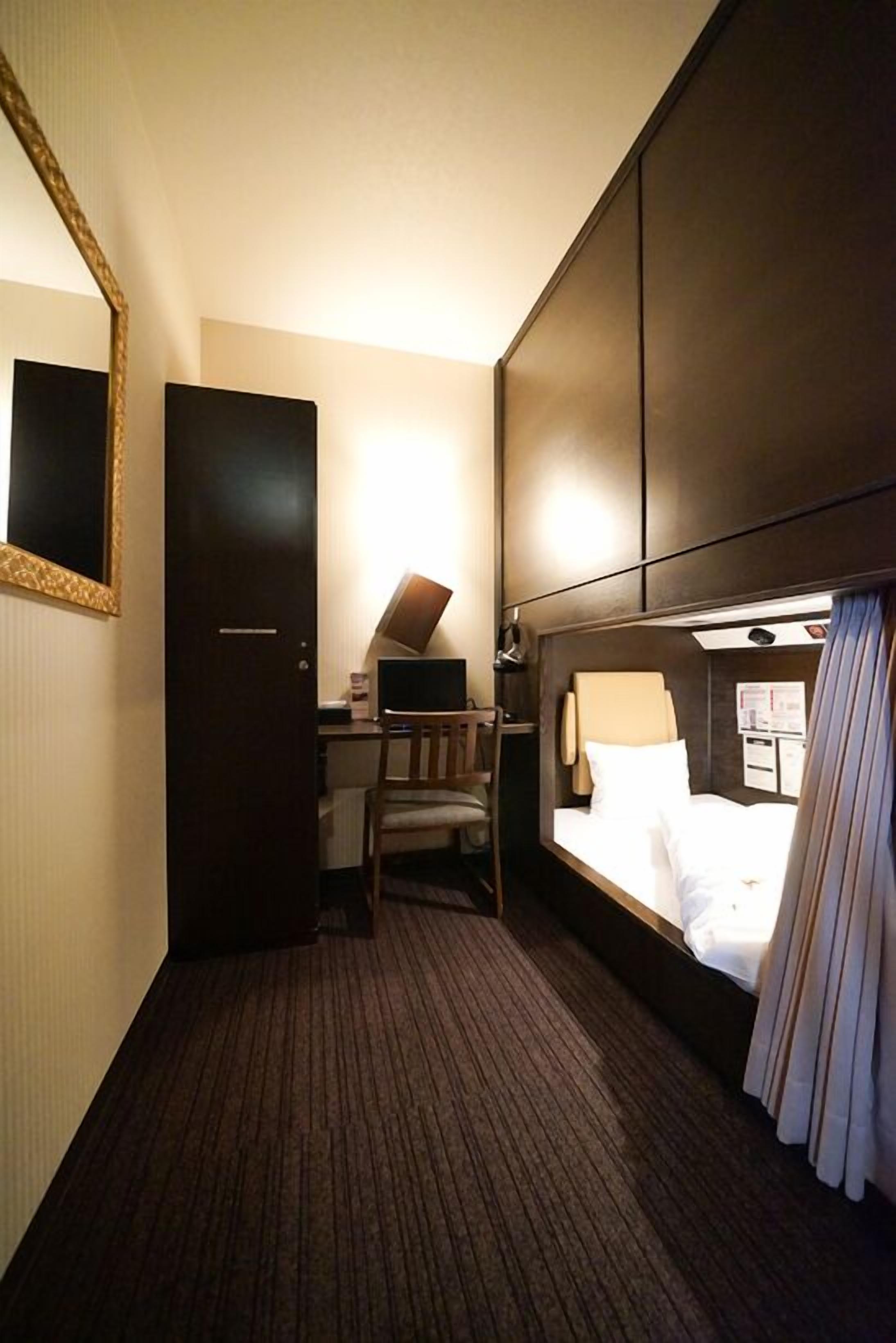Habitació d'hotel senzilla amb llit, escriptori, cadira, mirall i armari.