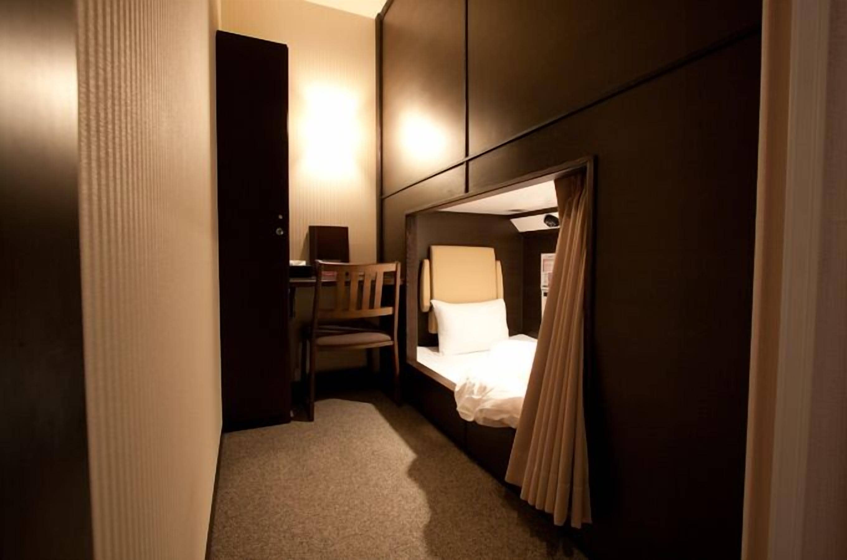 Habitació d'hotel petita amb un llit individual, una cadira, escriptori i un armari fixat a la paret.
