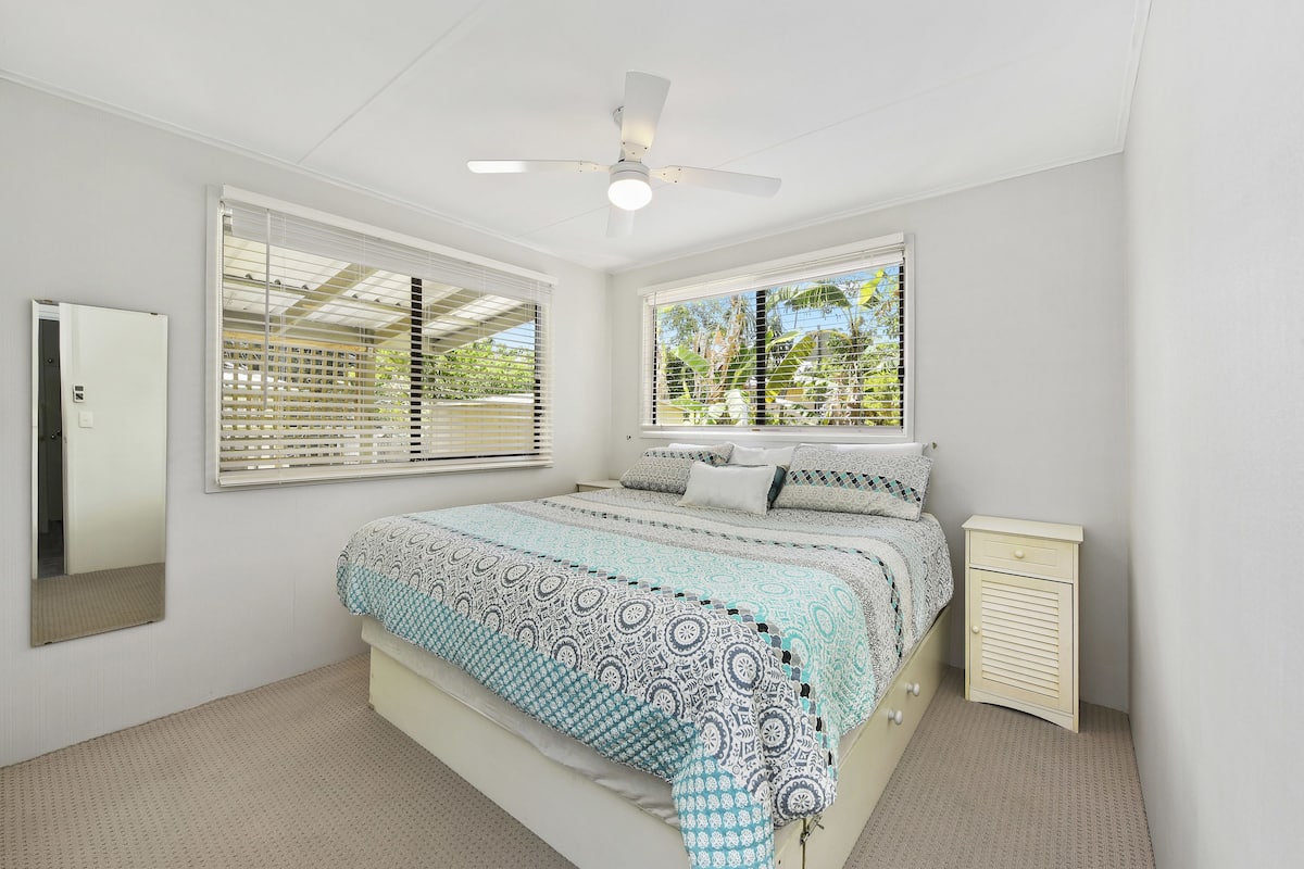 A bedroom with a bed, a bedside table, a ceiling fan, and two windows with blinds.