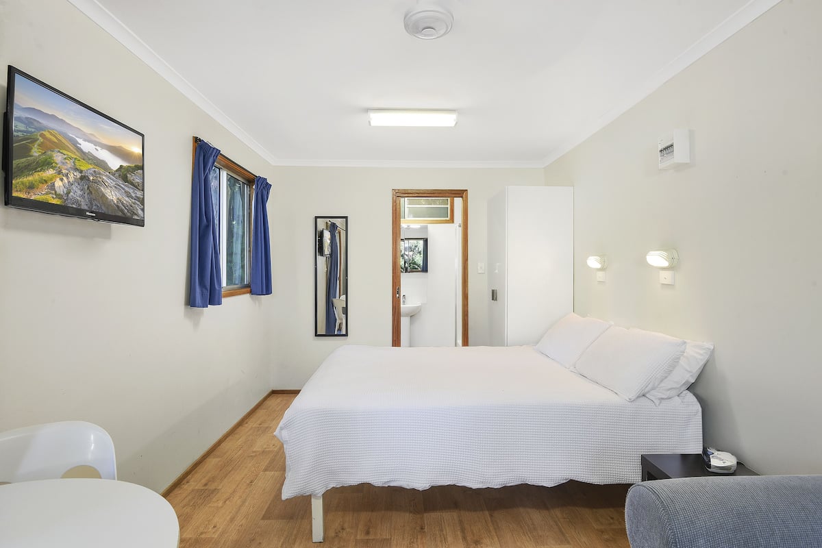 A bedroom with a bed, a desk, a chair, a TV, and a bathroom visible through an open door.