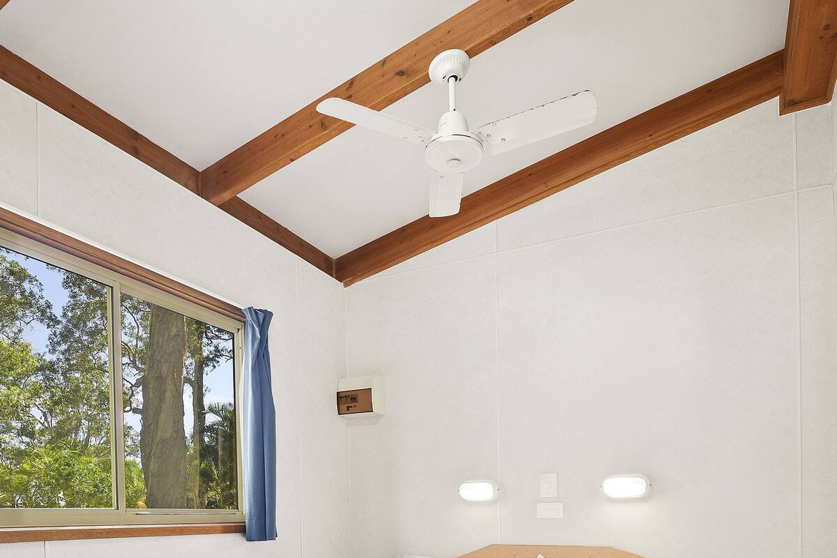 A bedroom with a bed, wooden beams, a ceiling fan, a window with curtains, and wall-mounted lights.