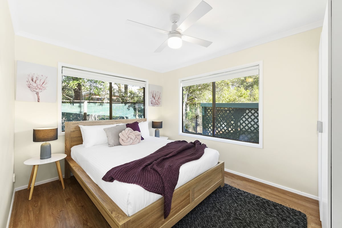A bedroom with a bed, two bedside tables, a ceiling fan, and two windows with views of greenery.