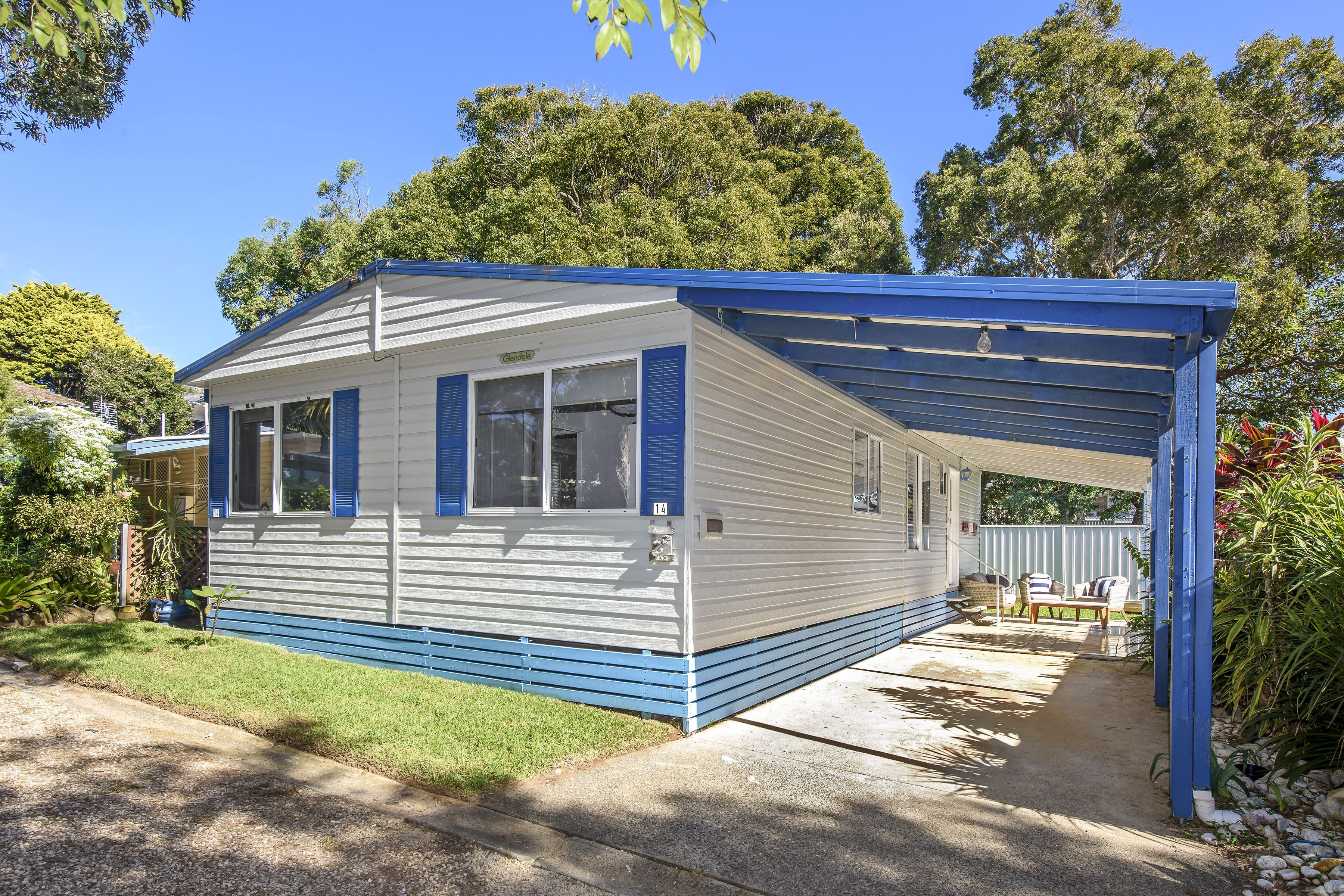 A single-story mobile home with a covered porch, a small garden, and a clear blue sky.