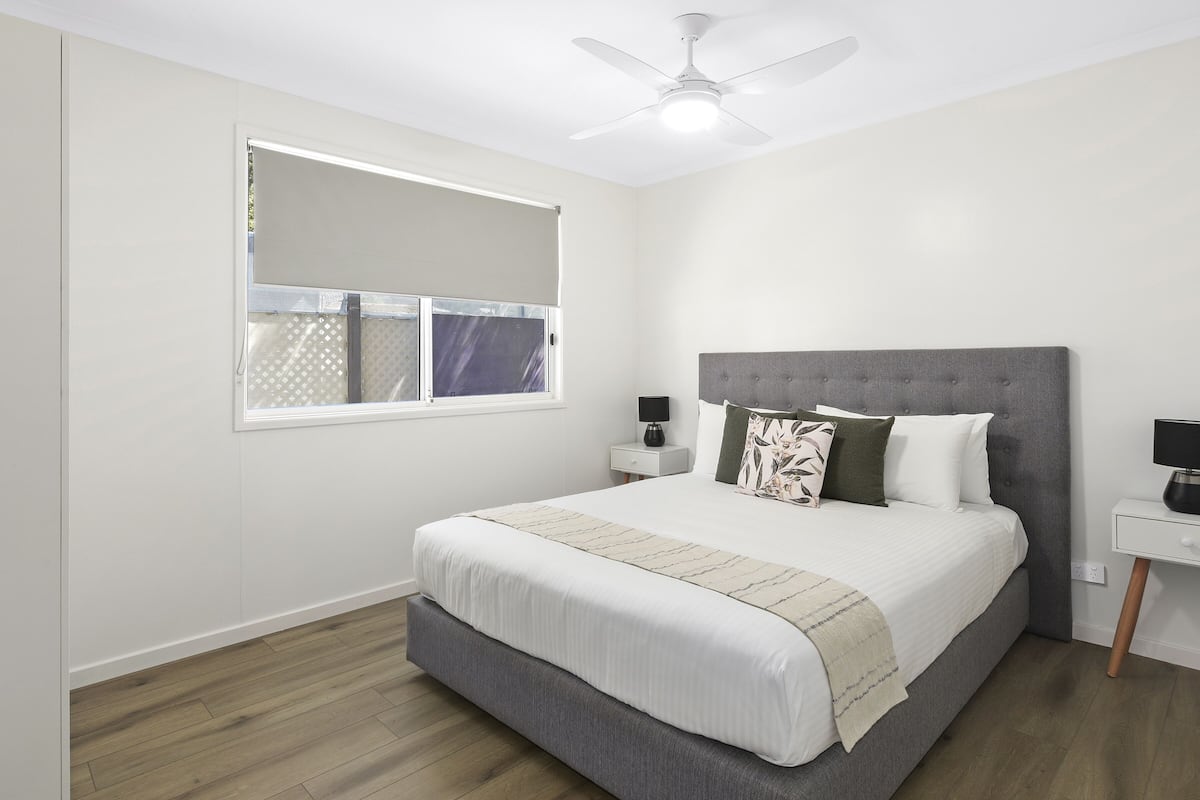 A bedroom with a bed, a nightstand, a lamp, a ceiling fan, and a window with blinds.