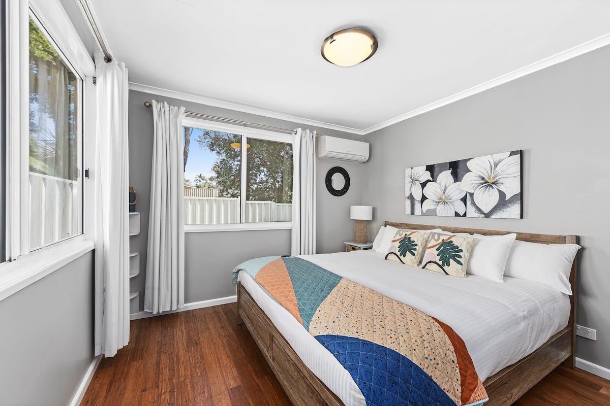A bedroom with a bed, a window with curtains, a wall-mounted air conditioner, a painting, and a wooden floor.