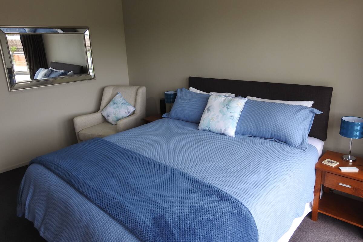 A neatly made bed with a blue bedspread and pillows, a chair with a cushion, a bedside table with a lamp, and a mirror on the wall.