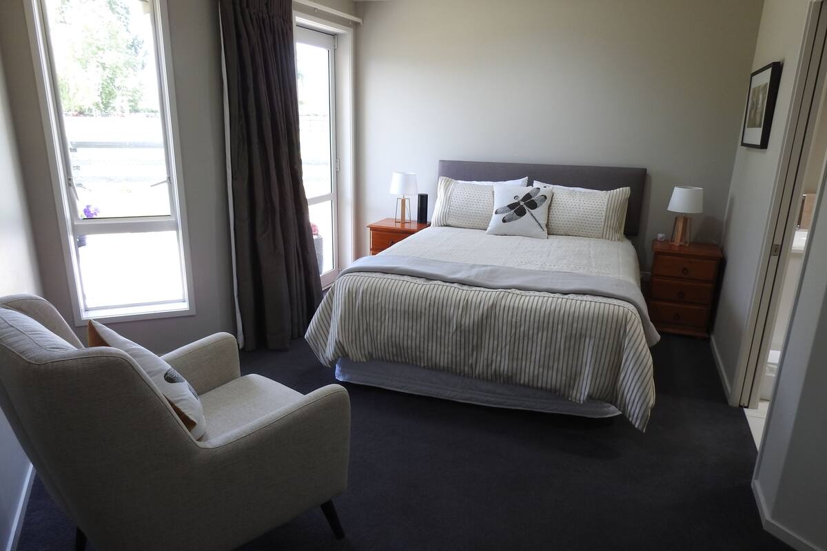A neatly arranged bedroom with a bed, a chair, a nightstand, and a window with curtains.