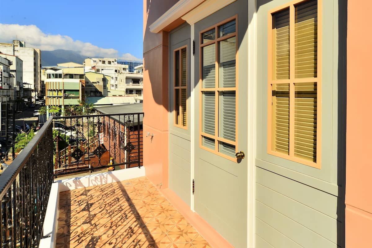 A balcony with a tiled floor and metal railing, overlooking a street with parked vehicles and buildings.