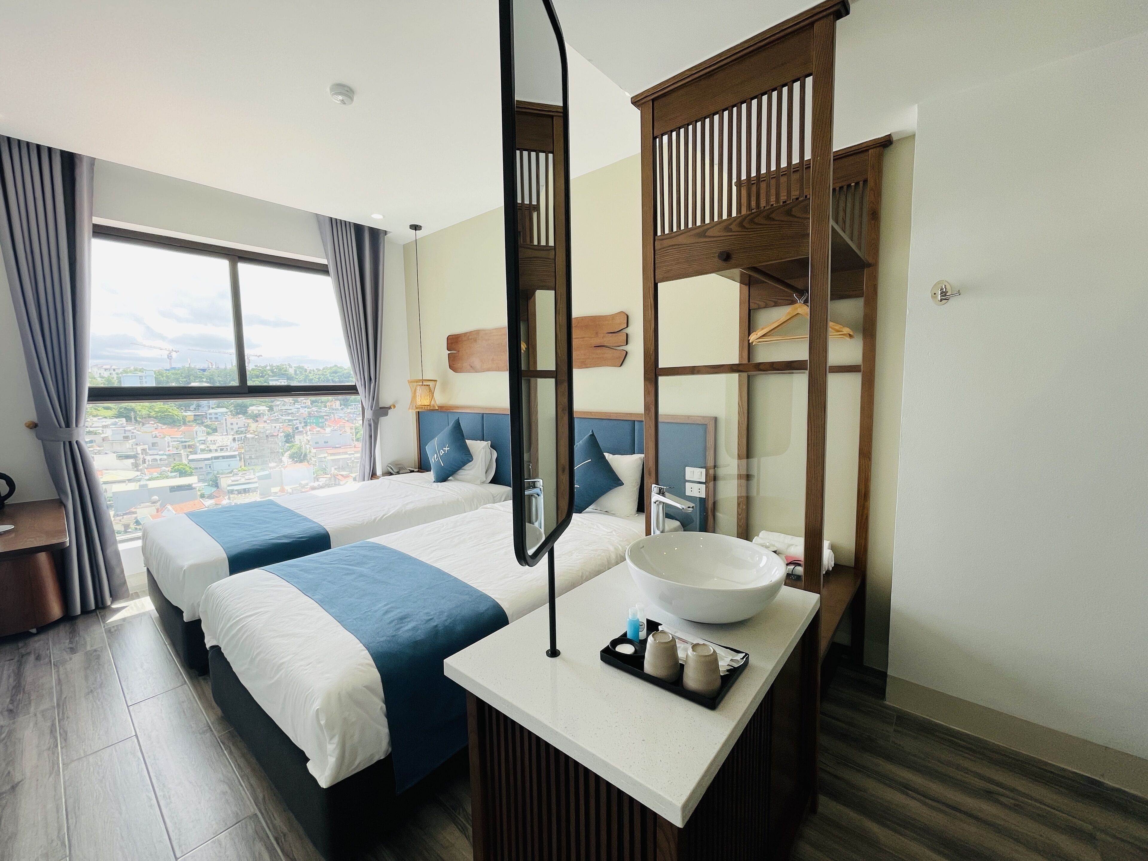 A hotel room with two beds, a large window with curtains, a wooden headboard, a mirror, a sink, and a tray with toiletries.