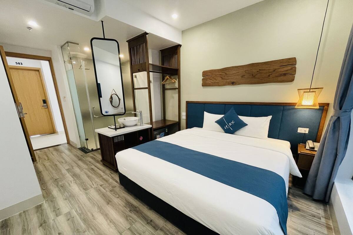 Habitación de hotel con una cama grande, una mesita de noche con lámpara, un espejo, un cabecero de madera y un armario.