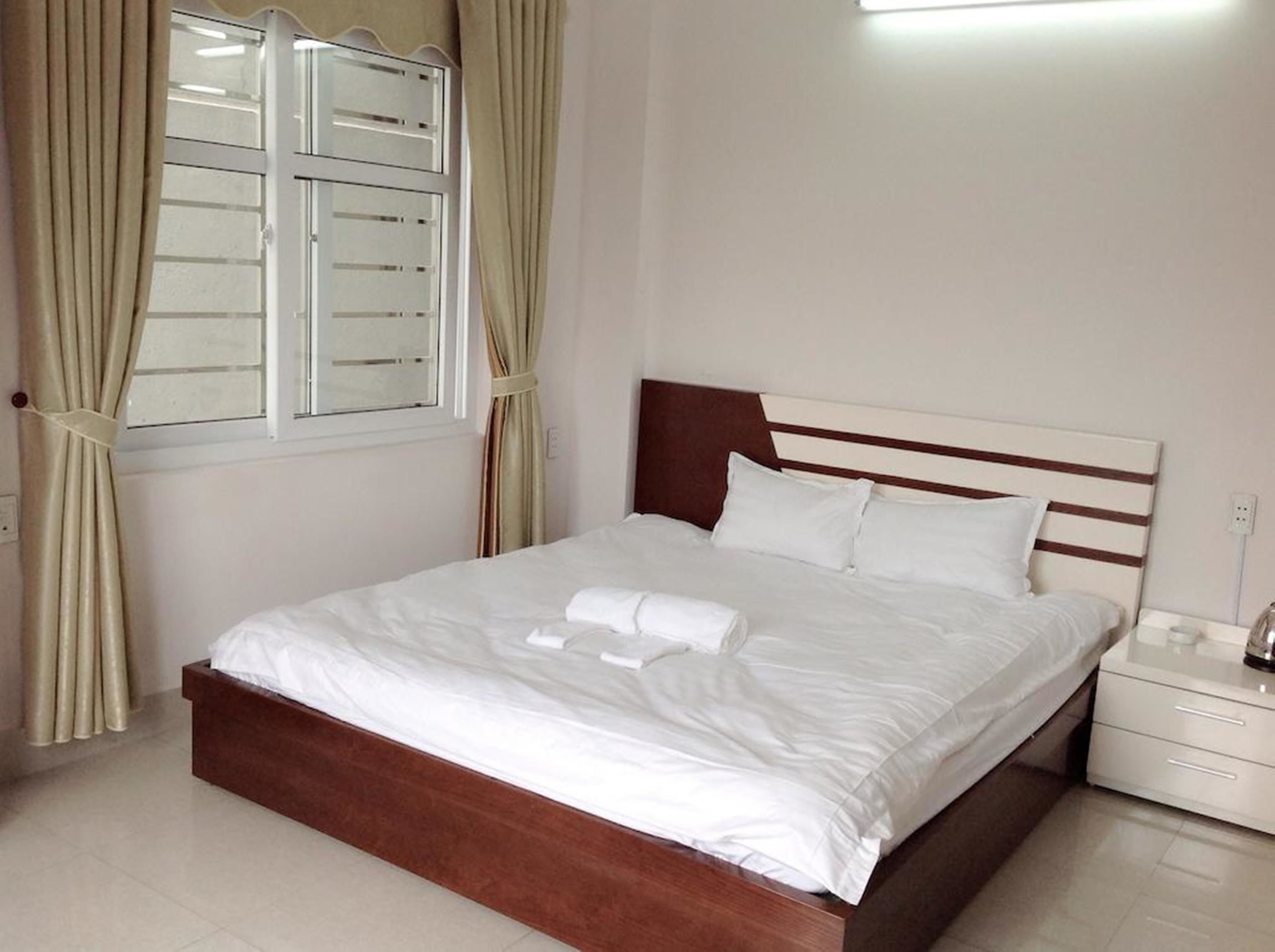 A neatly made bed with white linens and pillows, a wooden headboard, a bedside table with a lamp, and a window with curtains.