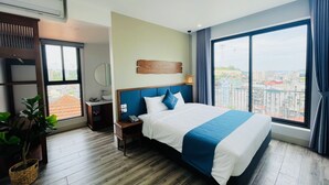 A hotel room with a large bed, a bedside table with a phone, a nightstand, a wall-mounted mirror, a potted plant, and a view of the city through large windows. - The Light Hotel (Bai Chay)