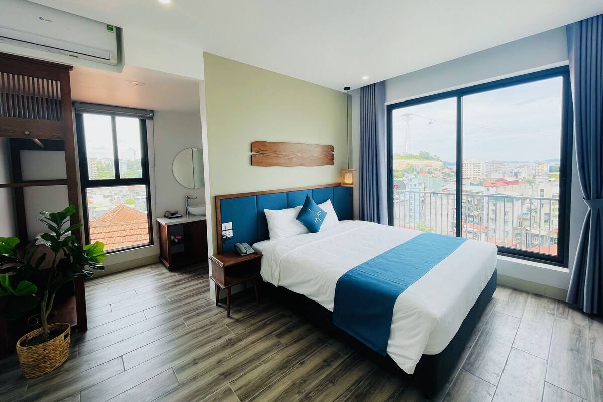 Un cuarto de hotel con una cama grande, una mesita de noche con un teléfono, una lámpara, un espejo en la pared, una planta en maceta y vistas de la ciudad a través de amplias ventanas.
