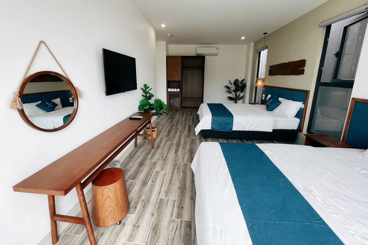 Habitación de hotel con dos camas, un escritorio de madera, un espejo redondo, un televisor y un ventanal amplio.