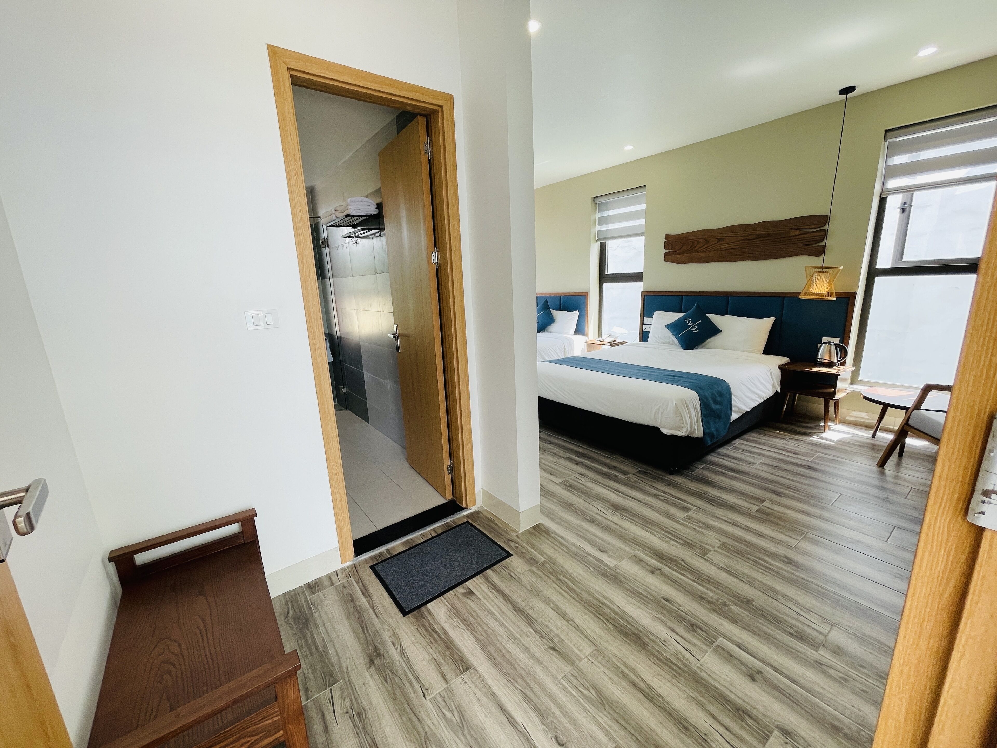 A hotel room with a wooden floor, a bed with blue bedding, a nightstand, a chair, and a window with blinds.