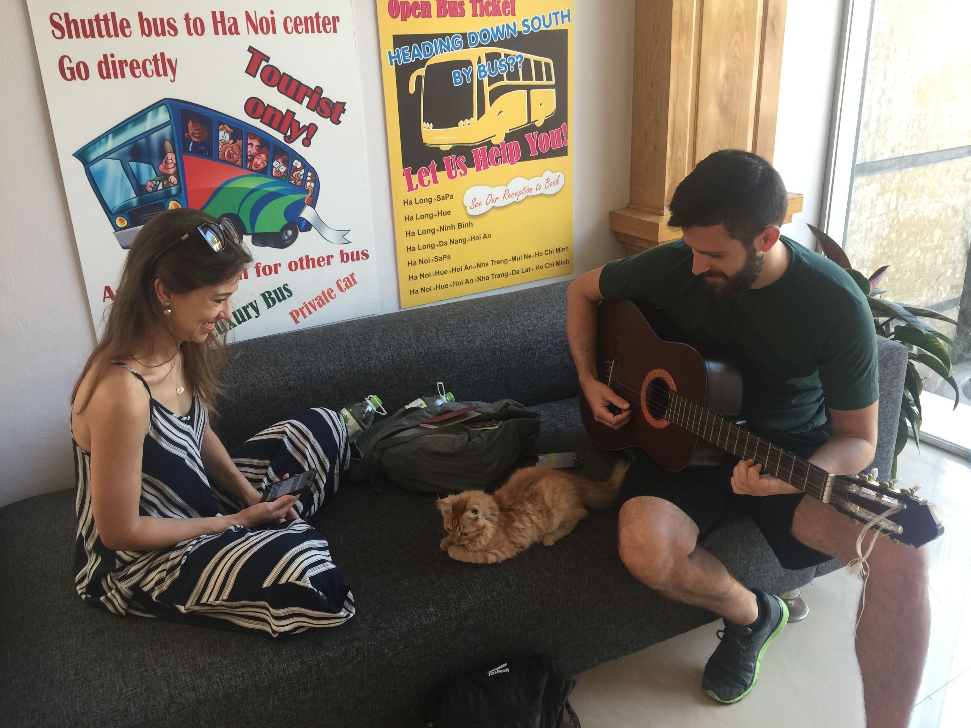 A man playing guitar and a woman sitting on a couch with a cat. There are posters on the wall advertising shuttle bus services.